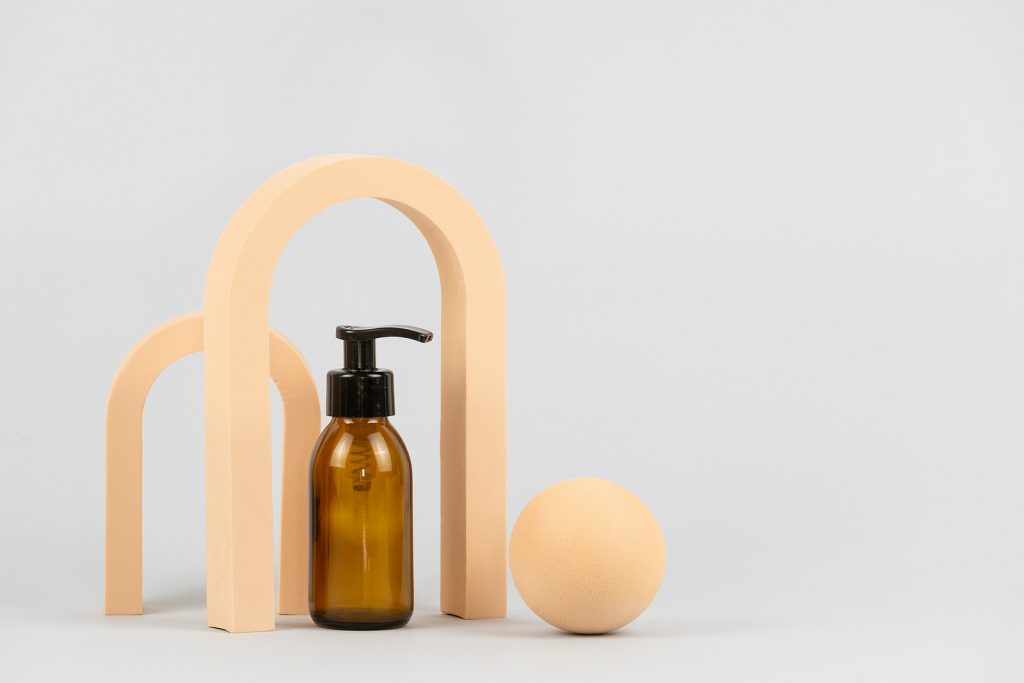 Dark amber glass bottle with body care product standing near geometric arch and ball on light gray background with copy space. Brand package mockup for skincare, haircare and beauty cosmetic ads