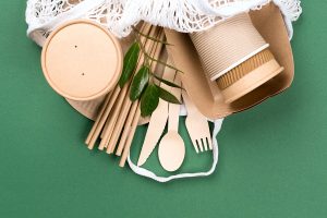 Paper utensils in white cotton net bag over green background.