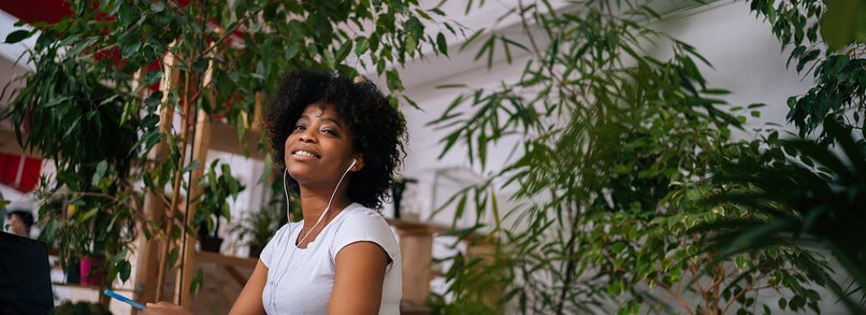 Low-angle portrait of pretty curly African American woman listening music in headphones sitting at table with laptop and smiling looking at camera, in light home office room with biophilic interior.