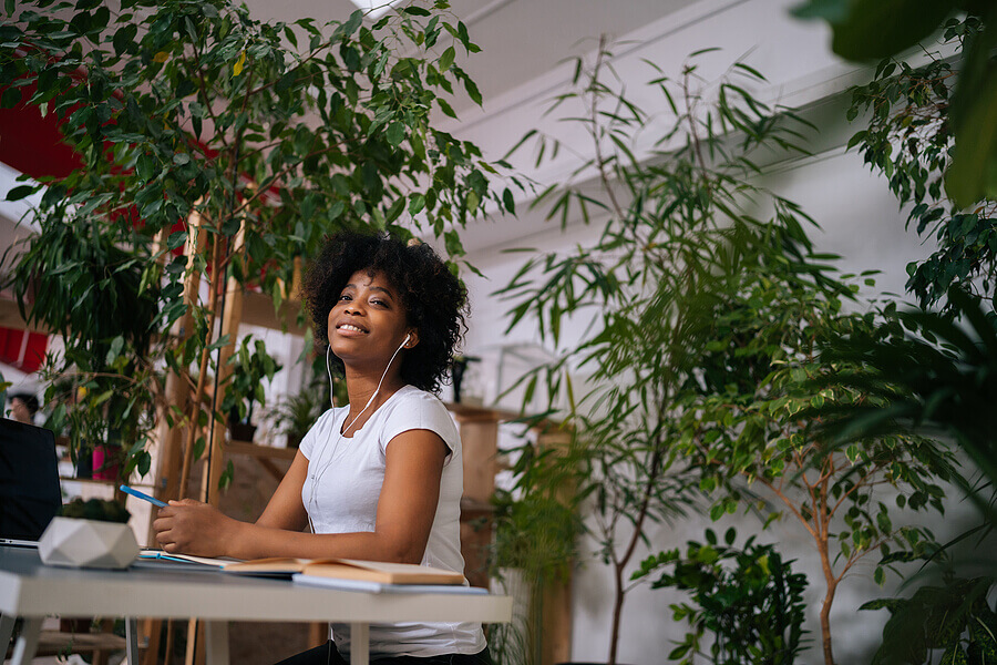 Low-angle portrait of pretty curly African American woman listening music in headphones sitting at table with laptop and smiling looking at camera, in light home office room with biophilic interior.