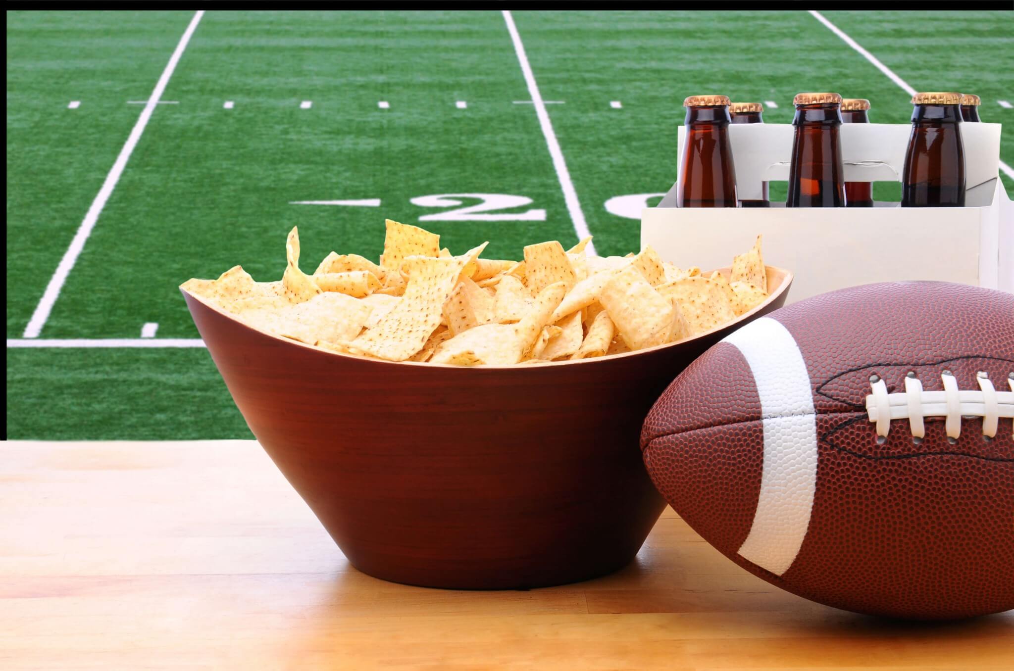 Chips, a six pack of beer, and an American football in front of the Super Bowl on TV.