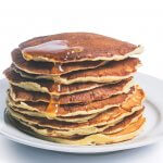 Pancakes stacked high on a white plate with honey drizzled over them.