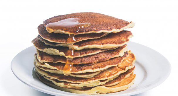 Pancakes stacked high on a white plate with honey drizzled over them.